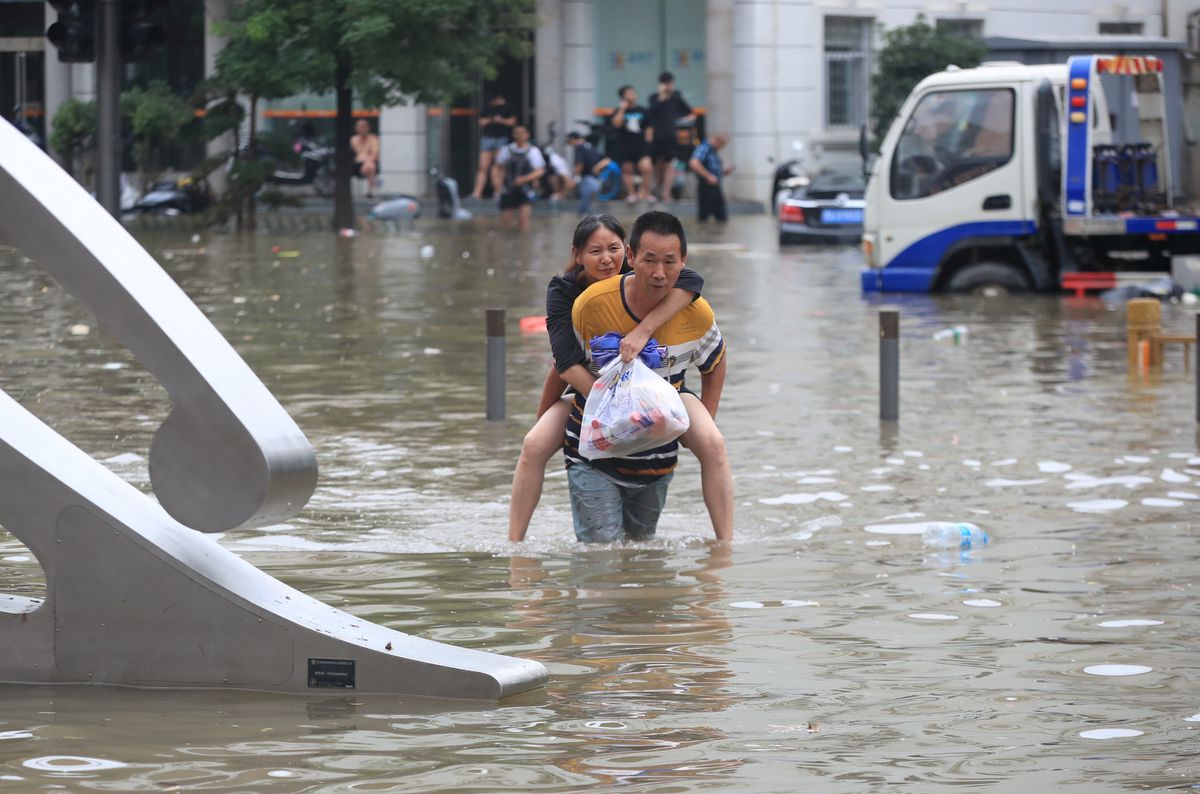 Một người đ&agrave;n &ocirc;ng c&otilde;ng một phụ nữ lội qua con đường ngập lụt sau trận mưa lớn ở Trịnh Ch&acirc;u ng&agrave;y 21/7 - Ảnh: China Daily/Reuters