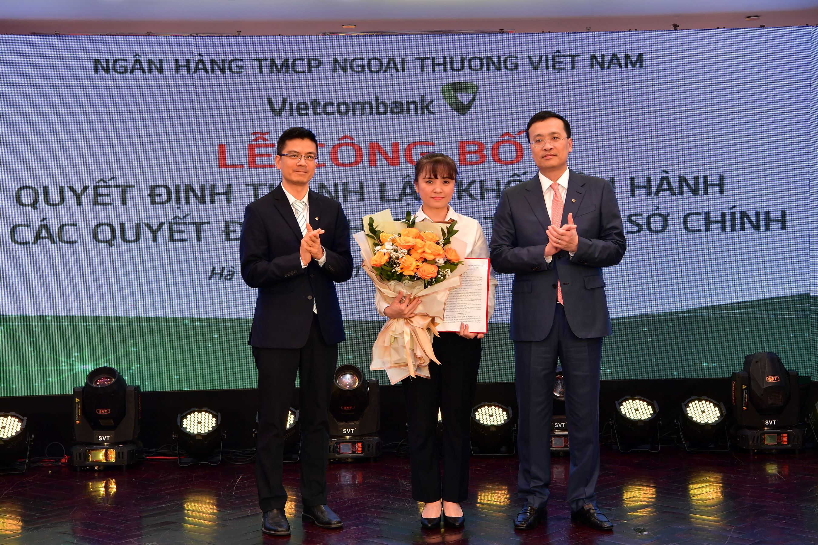 &Ocirc;ng Phạm Quang Dũng - Chủ tịch HĐQT (b&ecirc;n phải) trao quyết định v&agrave; &ocirc;ng Trần Thanh Nam -&nbsp;Gi&aacute;m đốc Khối Vận h&agrave;nh (b&ecirc;n trái) tặng hoa ch&uacute;c mừng b&agrave; Th&aacute;i Thị Thu Hằng - t&acirc;n Ph&oacute; trưởng ph&ograve;ng phụ tr&aacute;ch Ph&ograve;ng Quản l&yacute; chất lượng -&nbsp;Trụ sở ch&iacute;nh Vietcombank.
